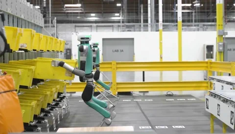 Az Agility Digit robotjának előző generációja a jellegzetes, hátrafelé hajló lábaival, miközben ládákat mozgat egy raktárban.