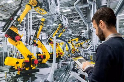 Egy mérnök tabletet használ egy gyár irányítására, mely tele van sárga FANUC robotkarokkal.
